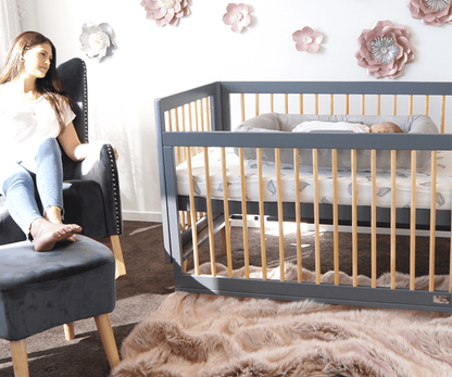 Woman sitting in a chair next to a baby hood cot and crib in a nursery with floral decorations on the wall.