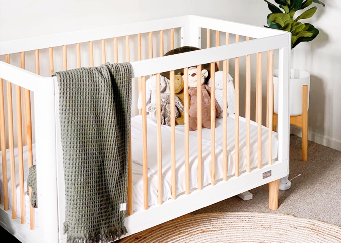 Babyhood White crib with wooden sides in a room with a plant and rug.